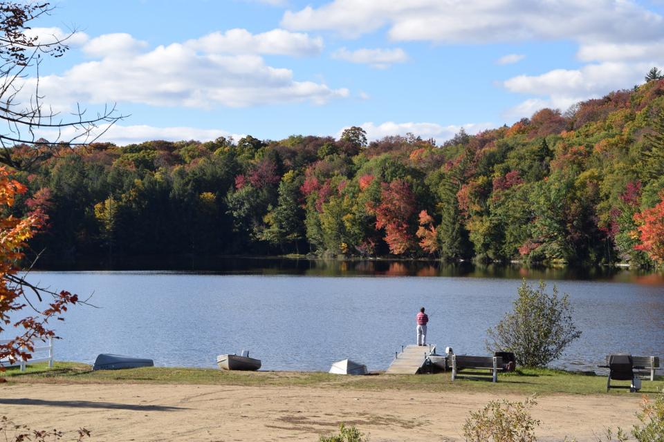 A single person stands fishing at the end of a short dock leading into a lake surrounded by fall foliage.