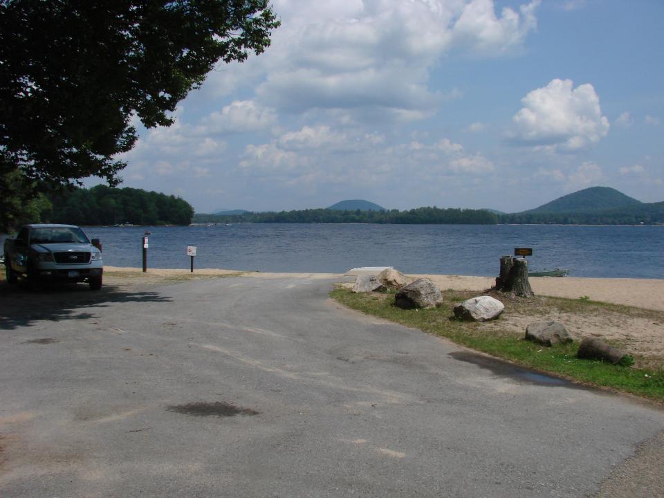 A paved drive leads to a flat, sandy beach on a blue lake with small, rounded mountains in the distance.