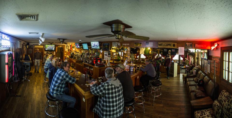 A wide, fisheye view of a rustic Adirondack bar. Men and women in plaid and flannel sit on stools.