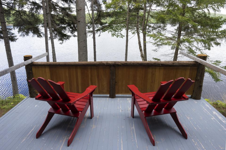 Two red Adirondack chairs on a small porch overlook a calm lake and trees.