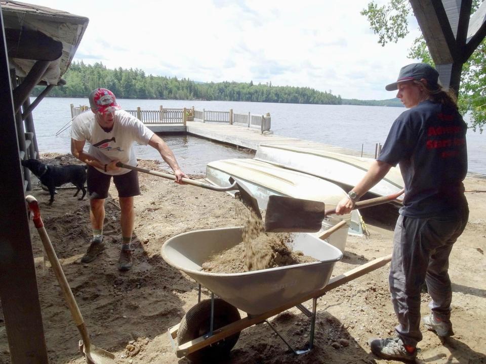 A man and woman shovel extra dirt away from a flooded boat house, in front of a lake.