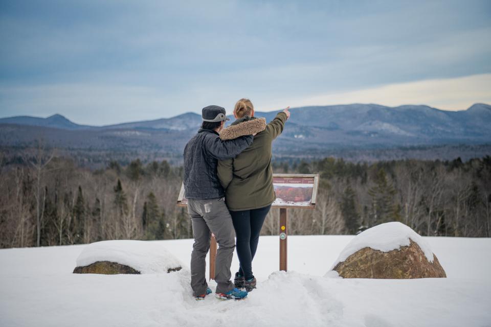 A couple wearing winter clothing looks toward snow covered mountains in the distance at the Route 30 Overlook in Indian Lake