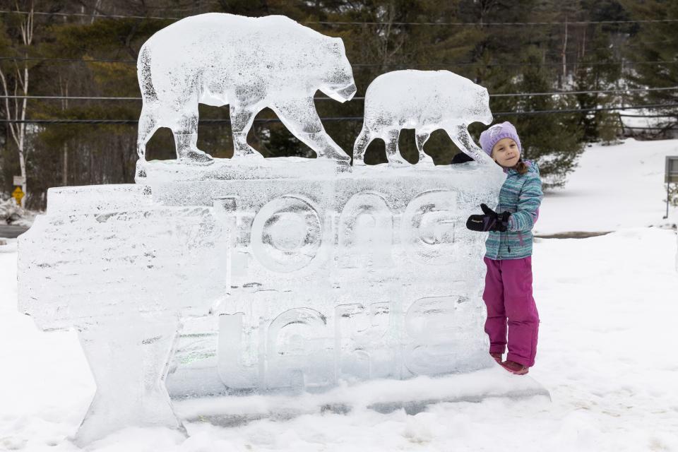 A girl in winter outdoor clothing hugs a large ice sculpture that shows a bear and reads "Long Lake."