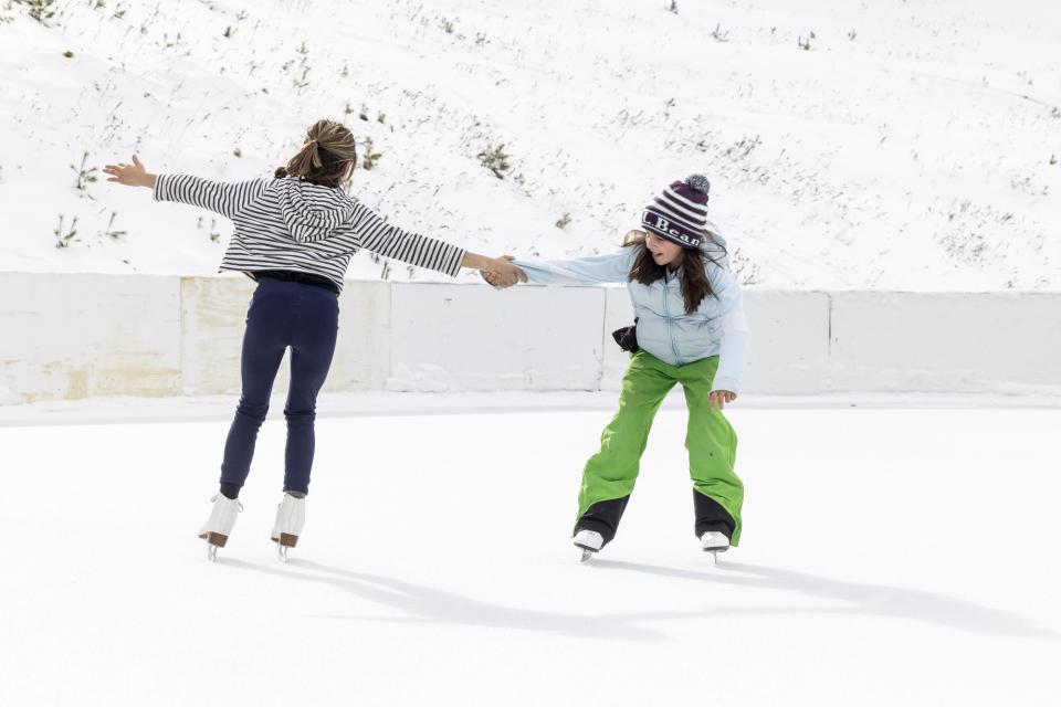 Two girls hold hands while ice skating on an outdoor rink.