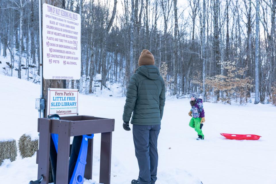 A woman stands next to a "library" of free sleds to borrow, while watching a child on a sled.