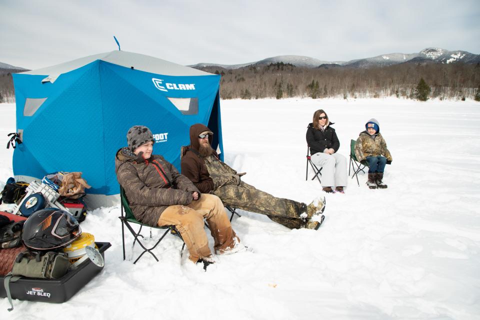 A family sits in snow suits around an ice fishing hole on a frozen lake.
