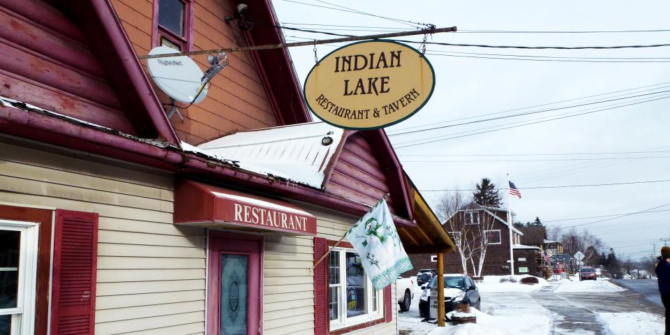 A sign with "Indian Lake Restaurant and Tavern" on it hanging off the outside of a restaurant in winter.