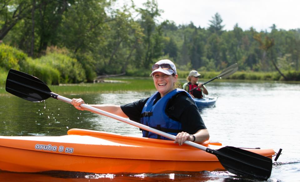 A woman in an orange kayak paddling in the lake wearing a black tshirt and blue life jacket with a baseball cap on and sunglasses on her cap smiling at the camera. in the background is a second kayaker