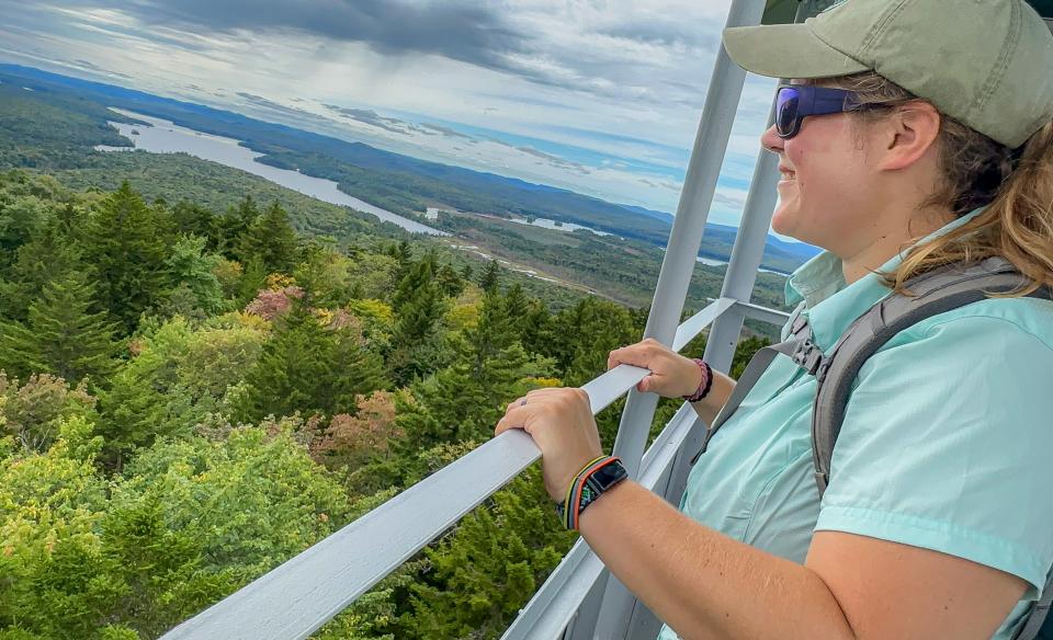 A hiker looks out of the fire tower on Buck Mountain over a forested landscape with a lake below.